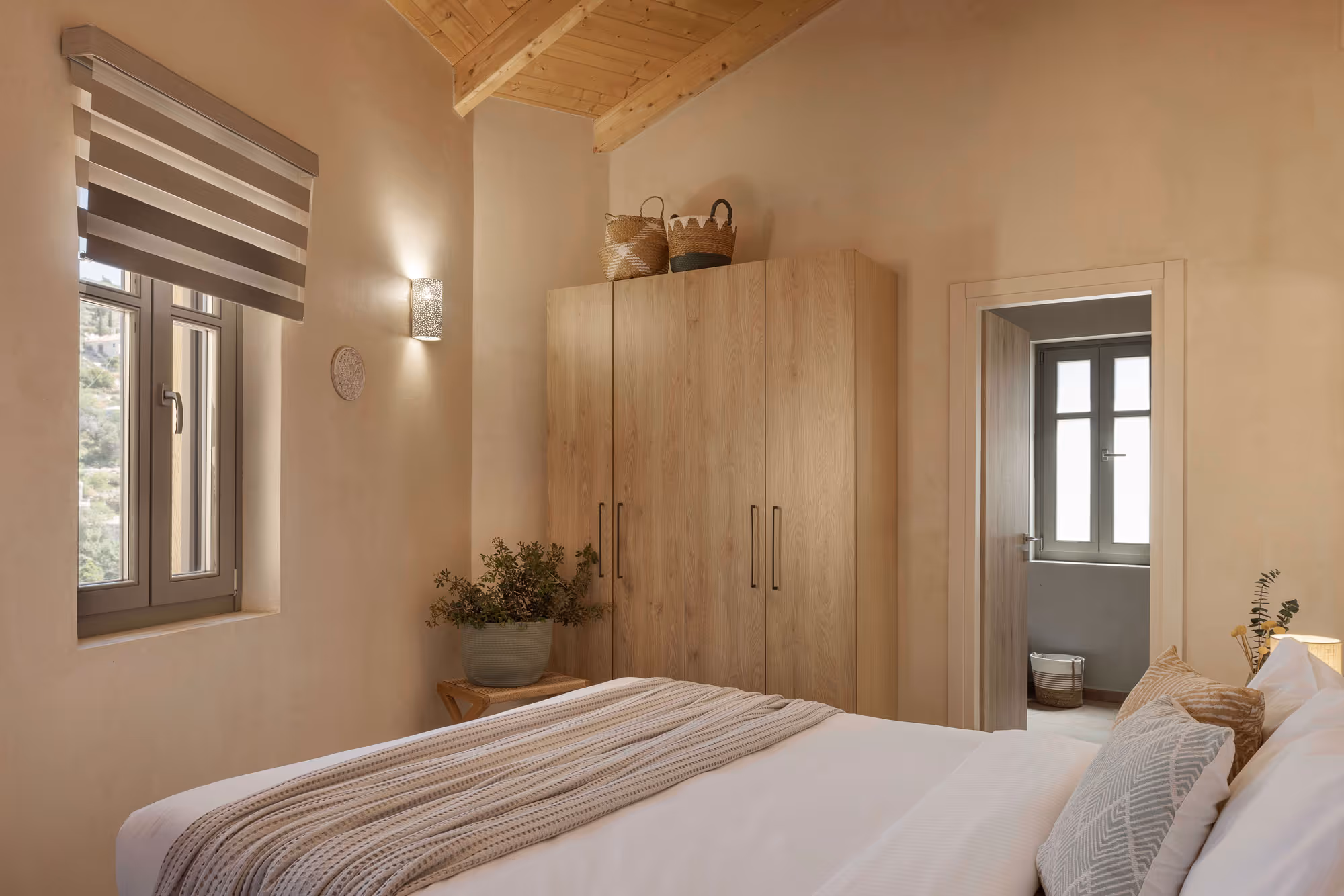 Cozy bedroom with wooden wardrobe, potted plant, striped window shade, and bed with pillows and blanket.