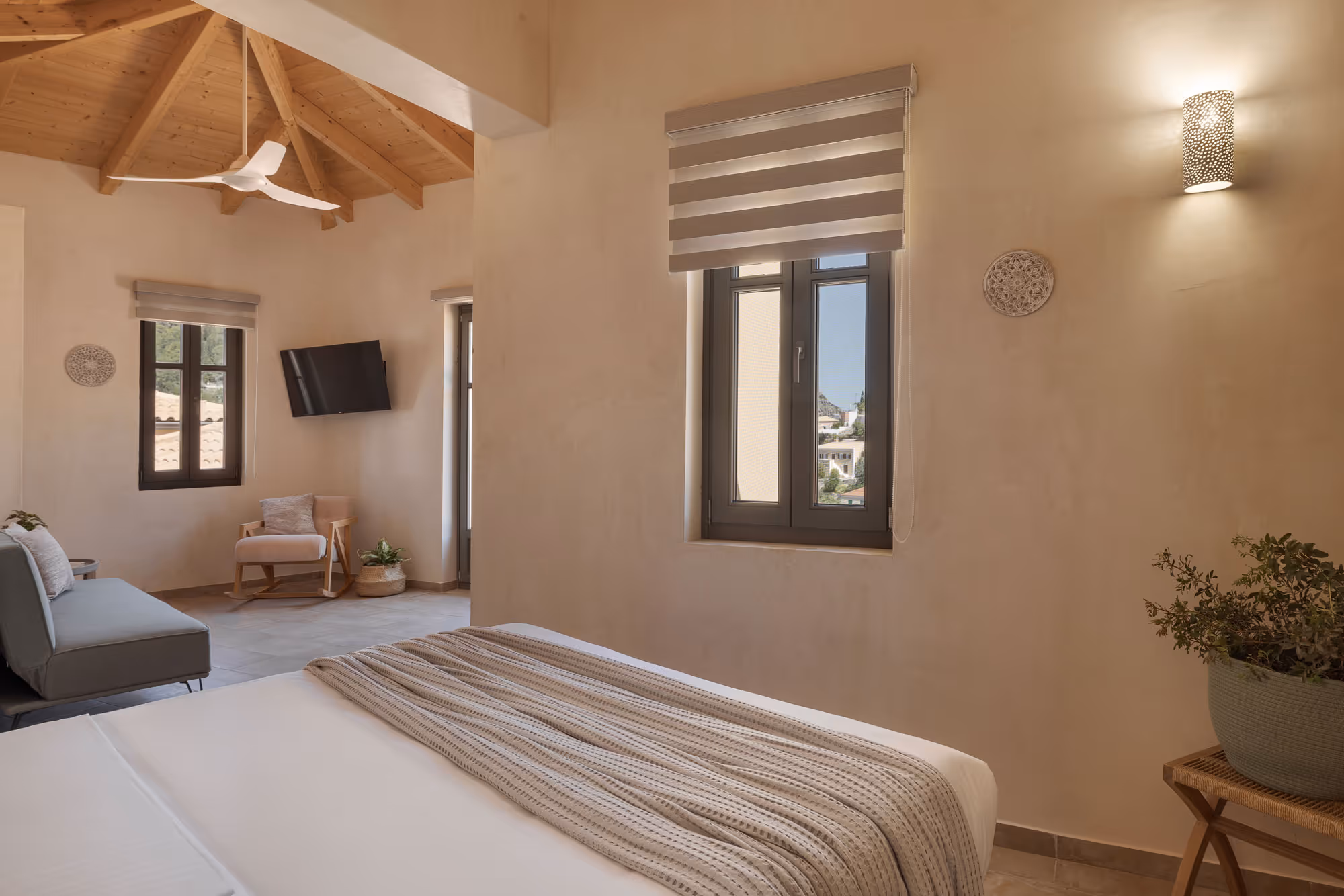 Cozy bedroom with wooden vaulted ceiling, white bed, striped blanket, window, chair, TV, and plants.