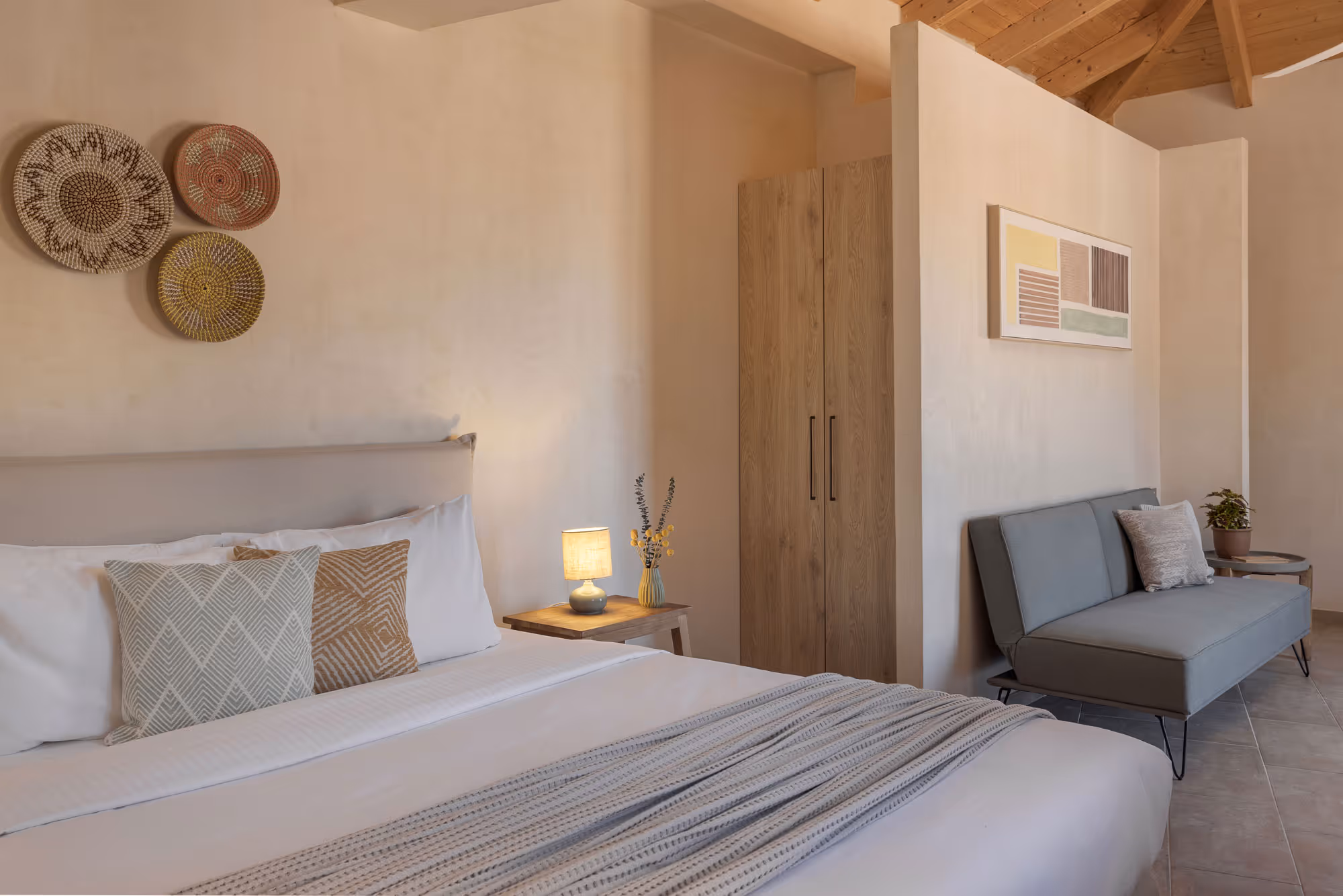 Cozy bedroom with a large bed, patterned pillows, a nightstand lamp, wardrobe, and gray sofa.