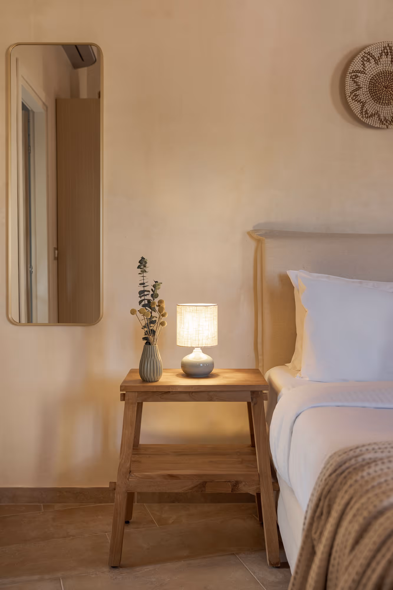 Cozy bedroom corner with a wooden nightstand, lit lamp, vase with dried flowers, and bed with white linens.