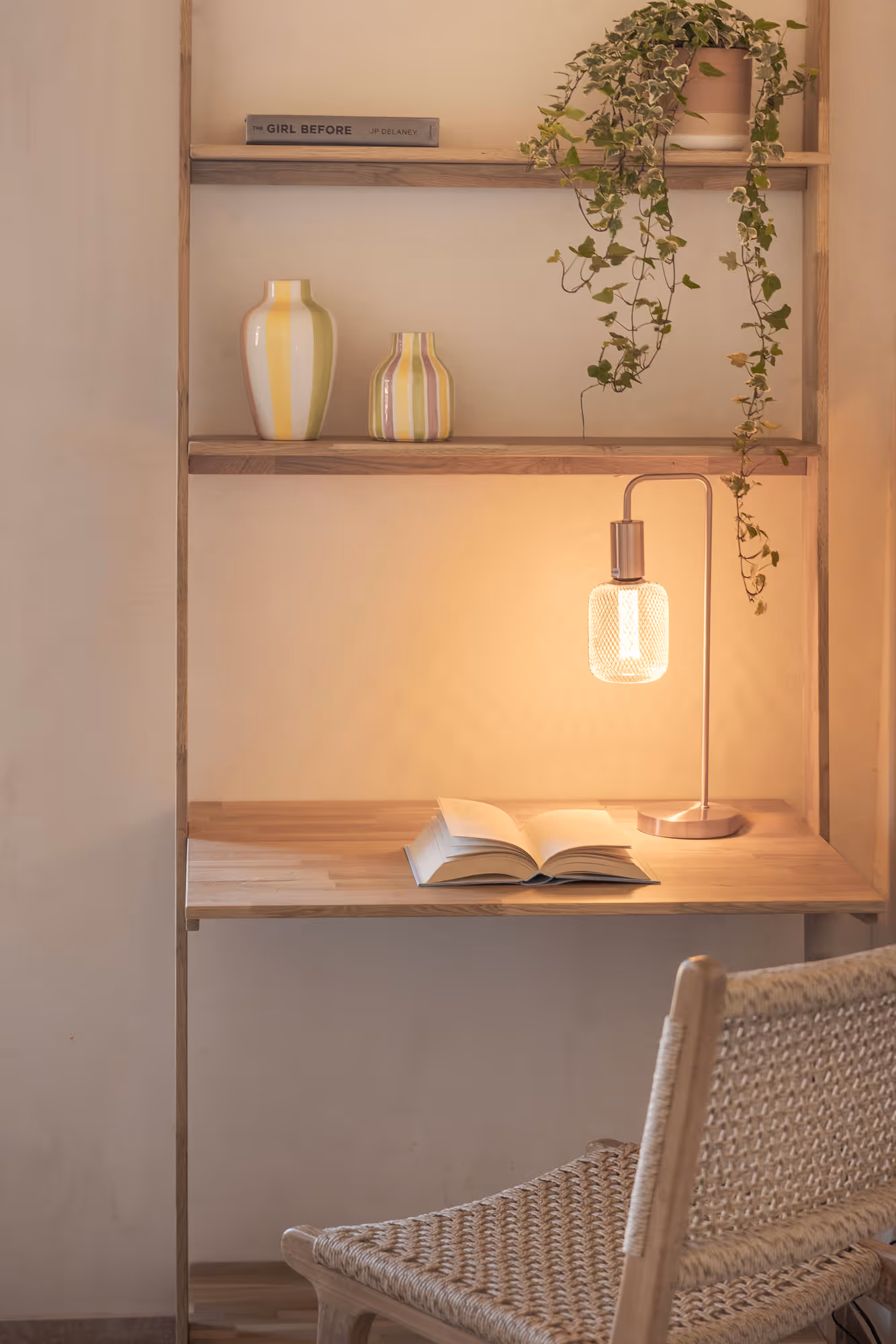 Cozy wooden shelf desk with lit lamp, open book, potted trailing plant, striped vases, and woven chair.