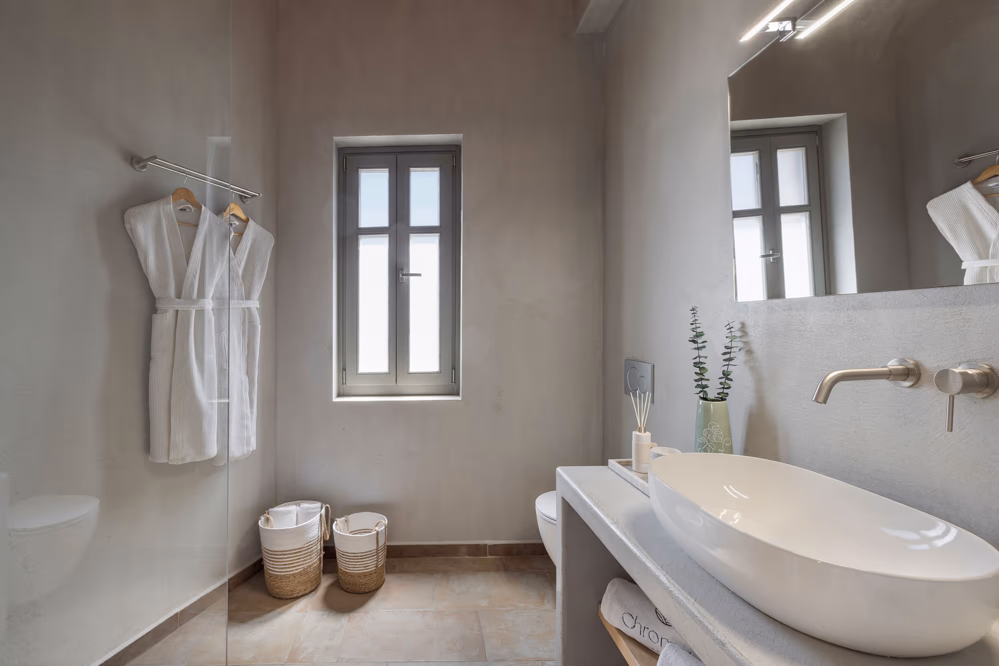 Minimalist bathroom with white oval sink, wall faucet, window, bathrobes, and woven baskets.