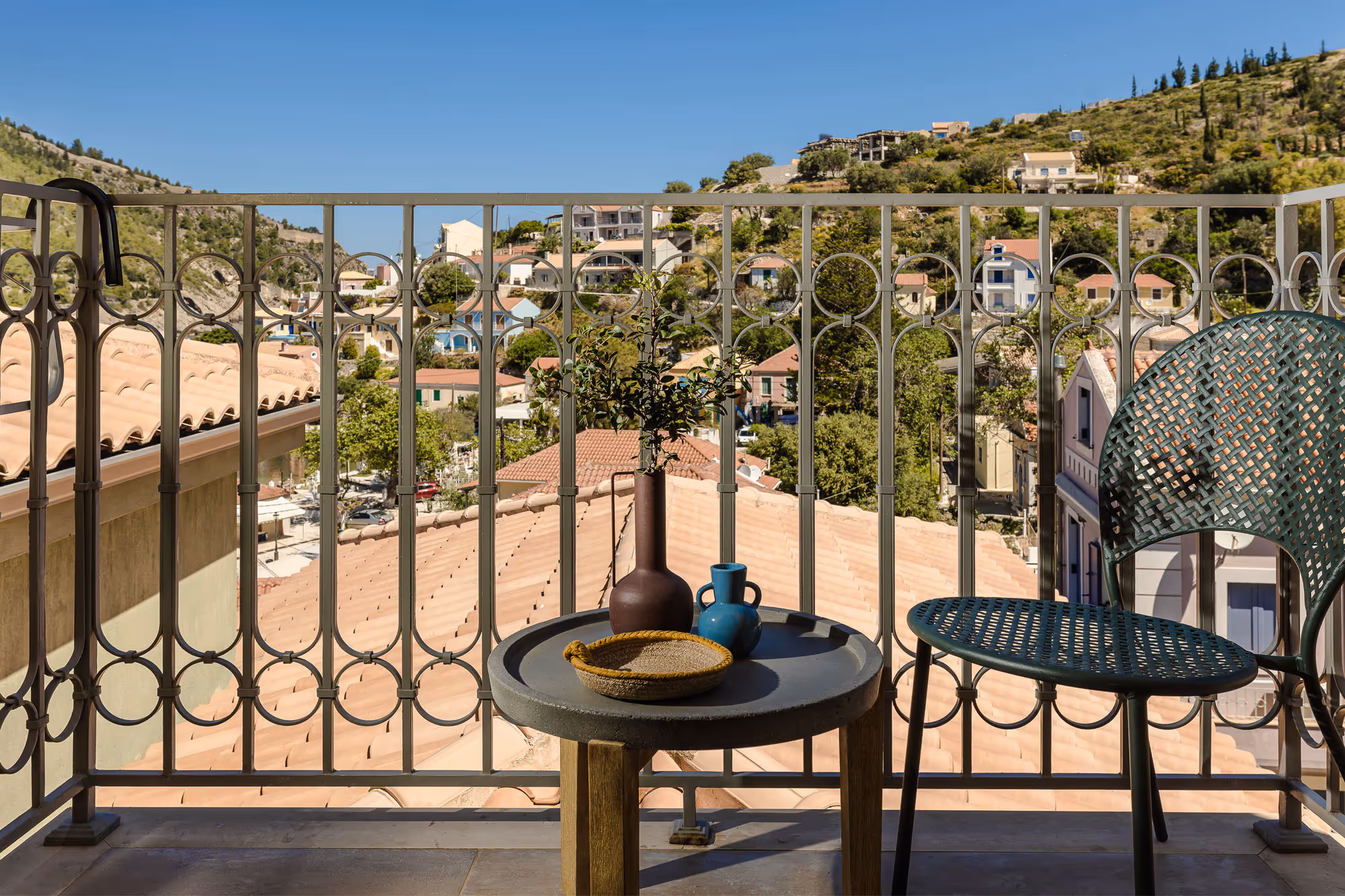 Balcony with round table holding two vases and basket, metal chair, overlooking hillside houses and blue sky.