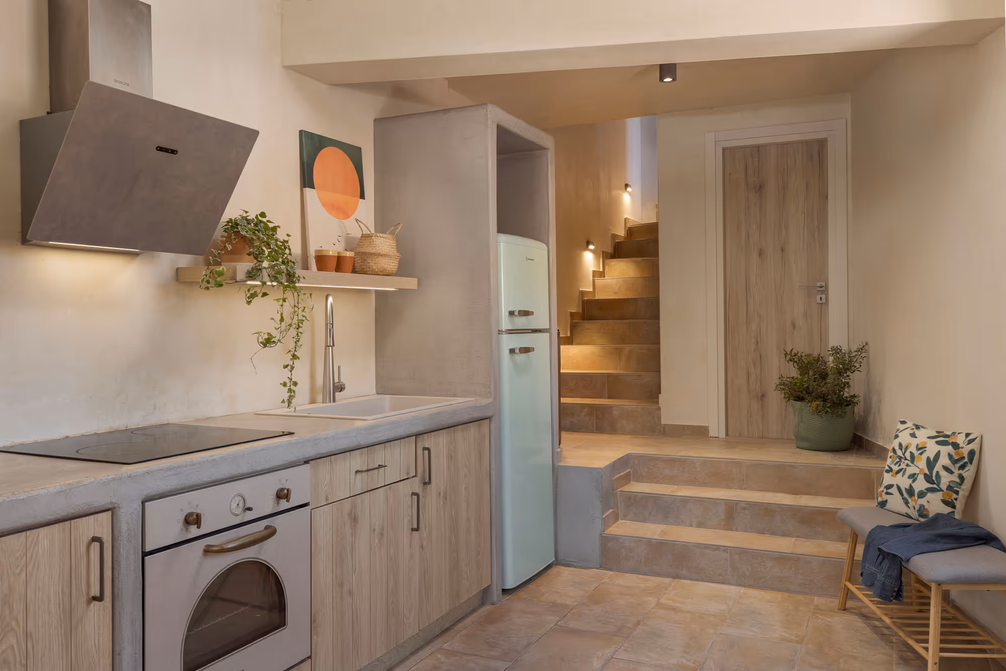 Modern kitchen with wooden cabinets, oven, pastel fridge, stairs, bench with pillow, and potted plants.