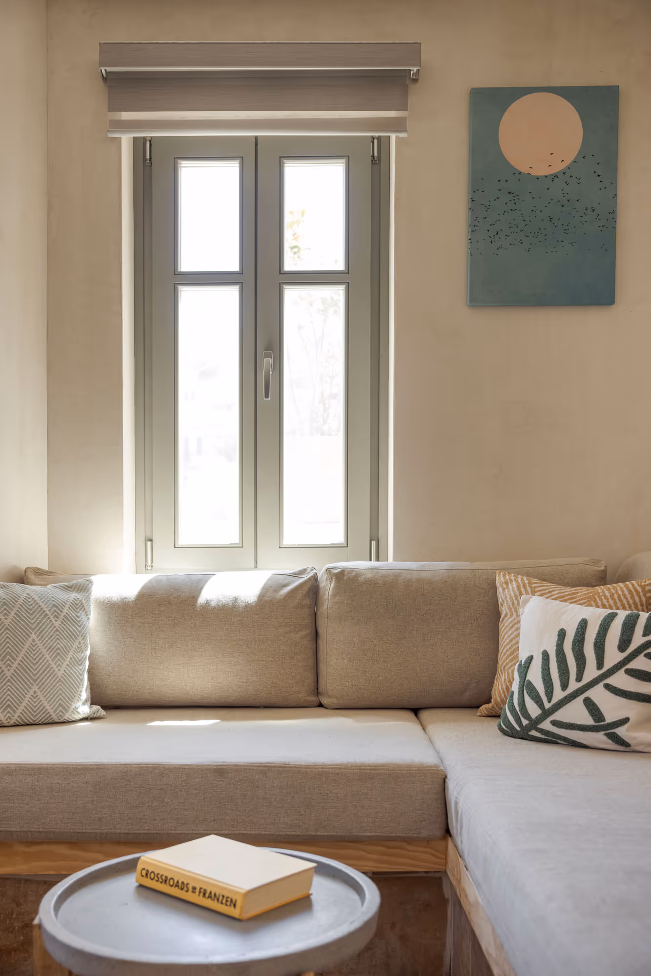 Bright living room with beige sectional sofa, patterned cushions, window, and a book on round table.