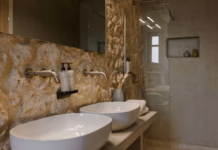 Modern bathroom with two white vessel sinks and wall-mounted faucets on textured stone wall.