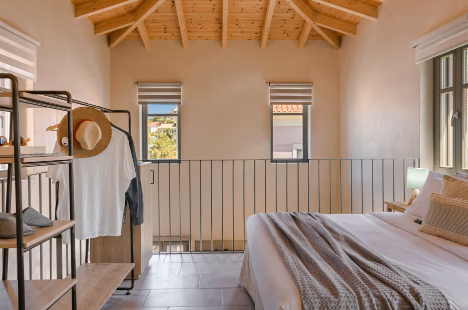 Bedroom loft with wooden ceiling, bed with pillows and blanket, and open clothes rack with hanging hat and shirt.