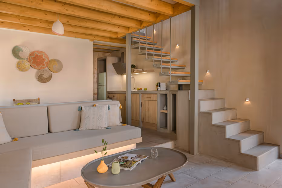 Cozy living room with beige sofa, round table, wooden ceiling, and staircase next to a small kitchen.