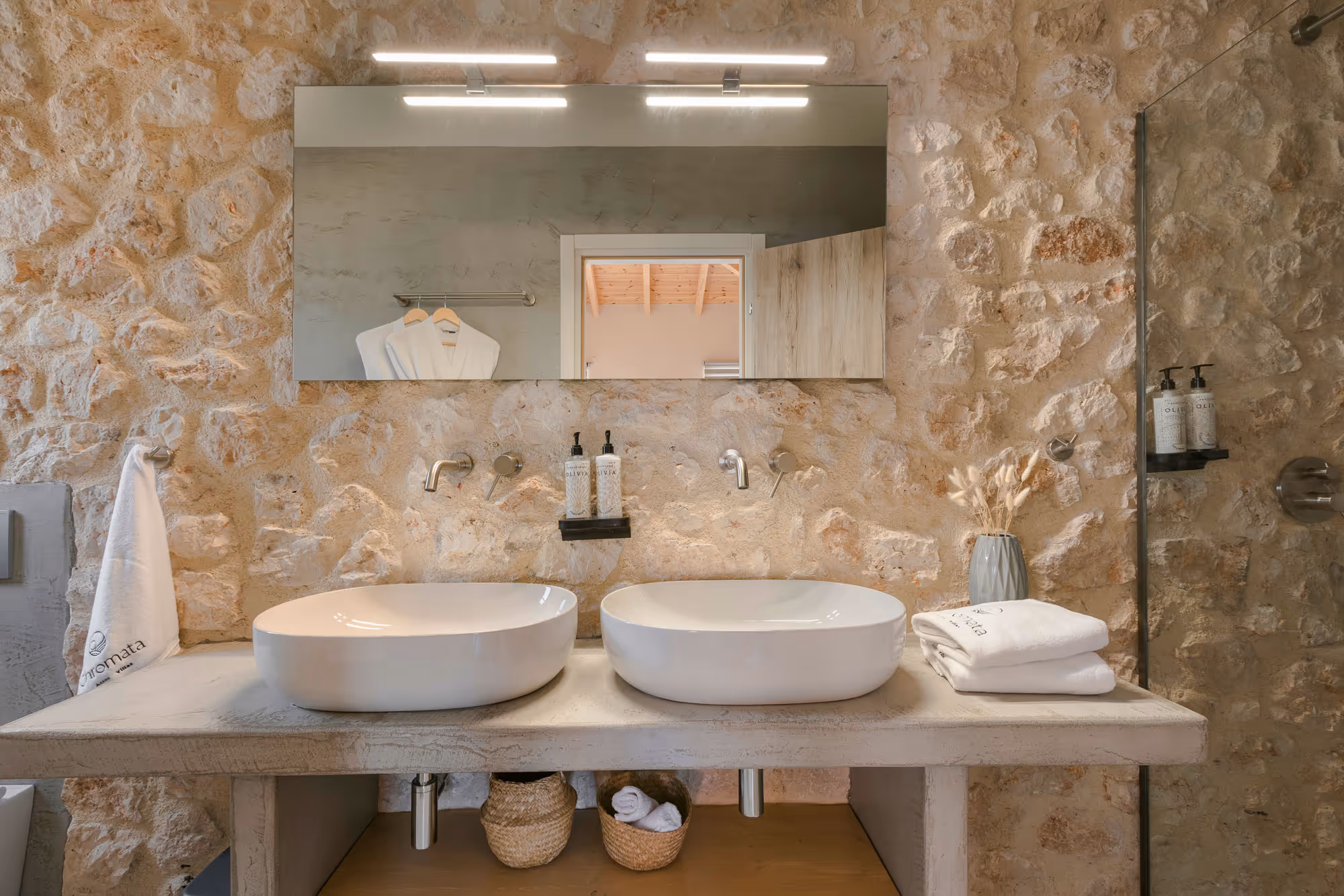 Rustic bathroom with stone wall, two white vessel sinks, large mirror, and folded towels.