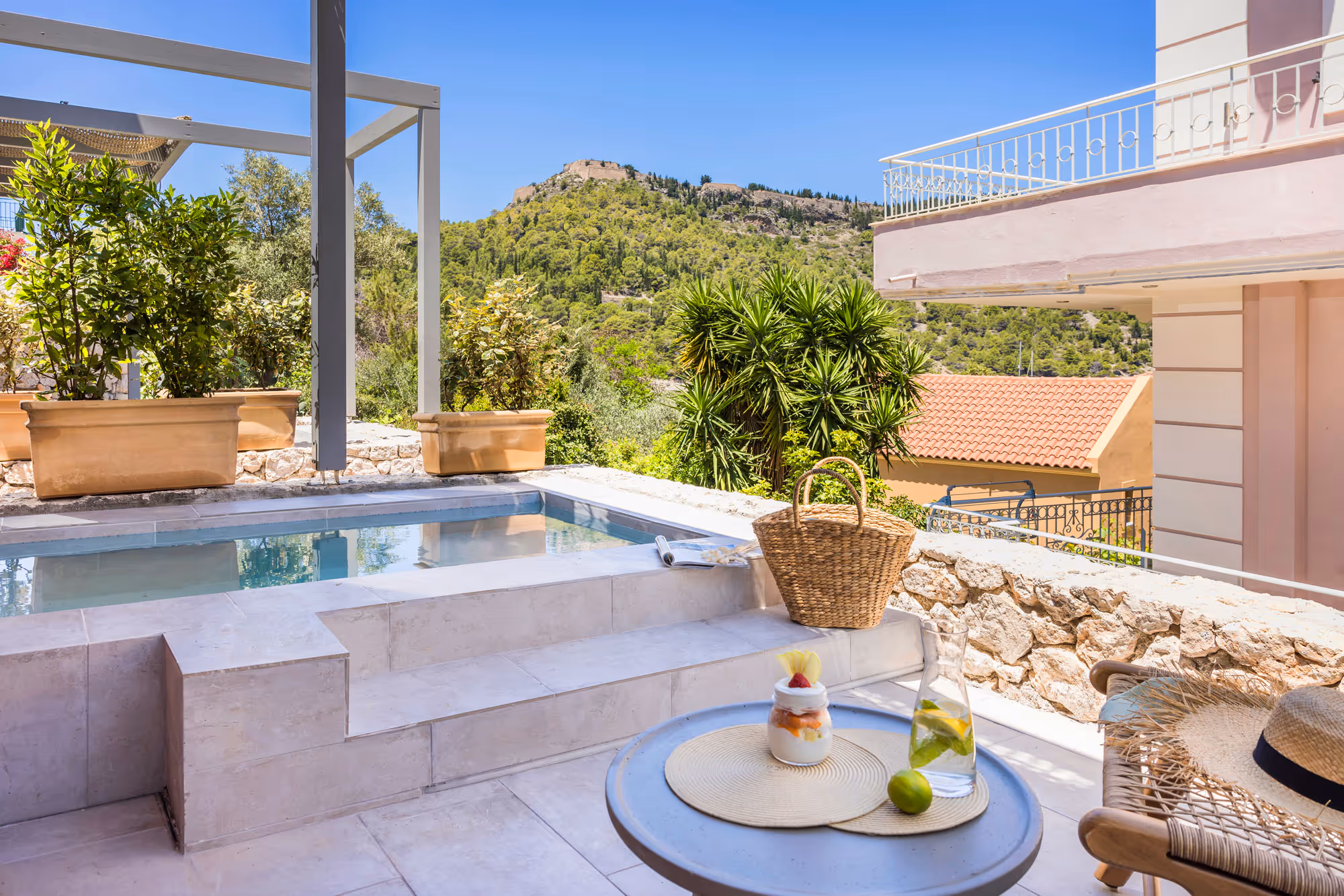 Sunny patio with pool, wicker basket, pitcher with lemon water, dessert, and mountain view.