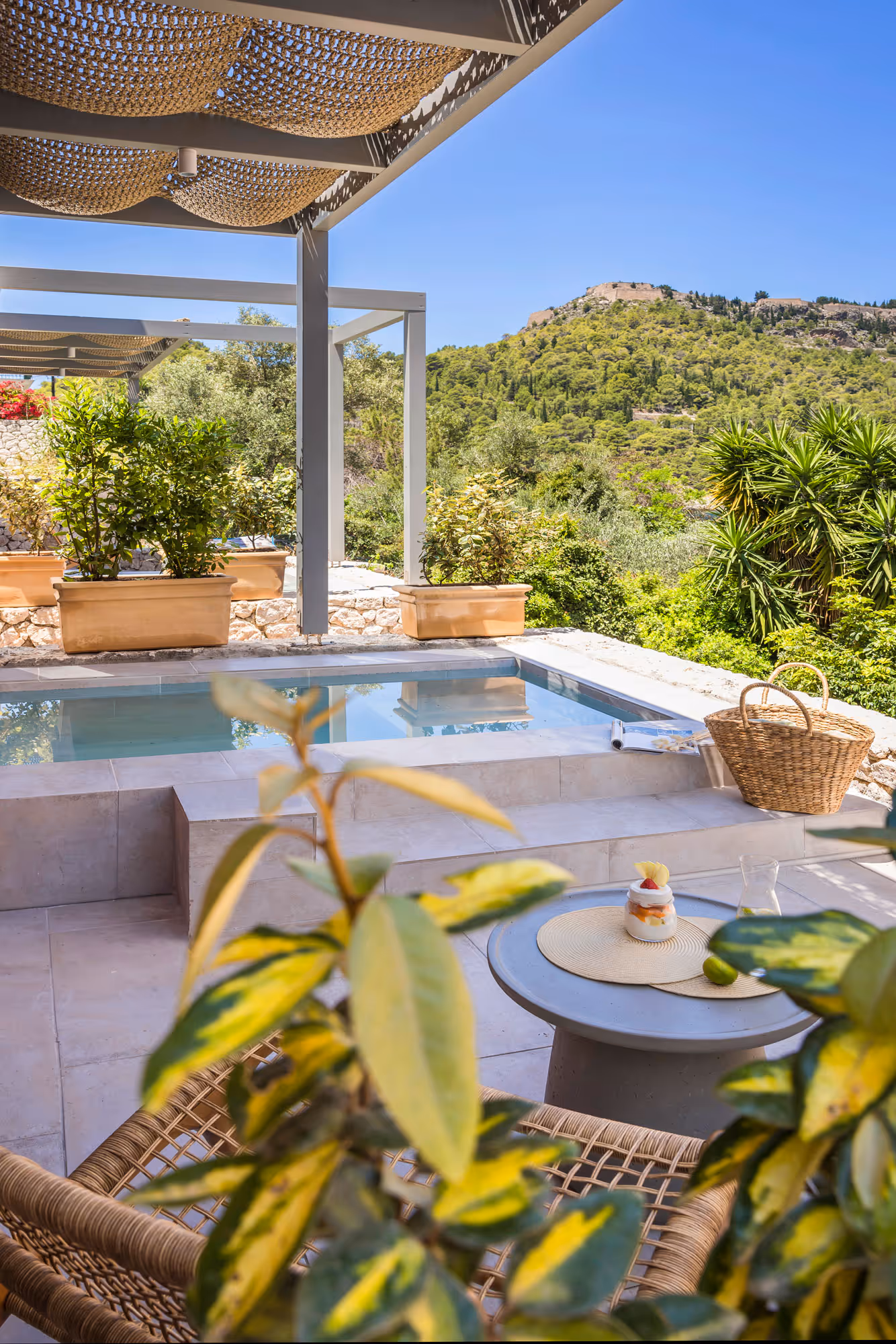 Outdoor patio with small pool, wicker chair, round table with dessert, and green hills in the background.